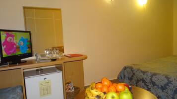 A hotel room with a television, a refrigerator, a desk with a phone, glasses, and a basket of fruit.