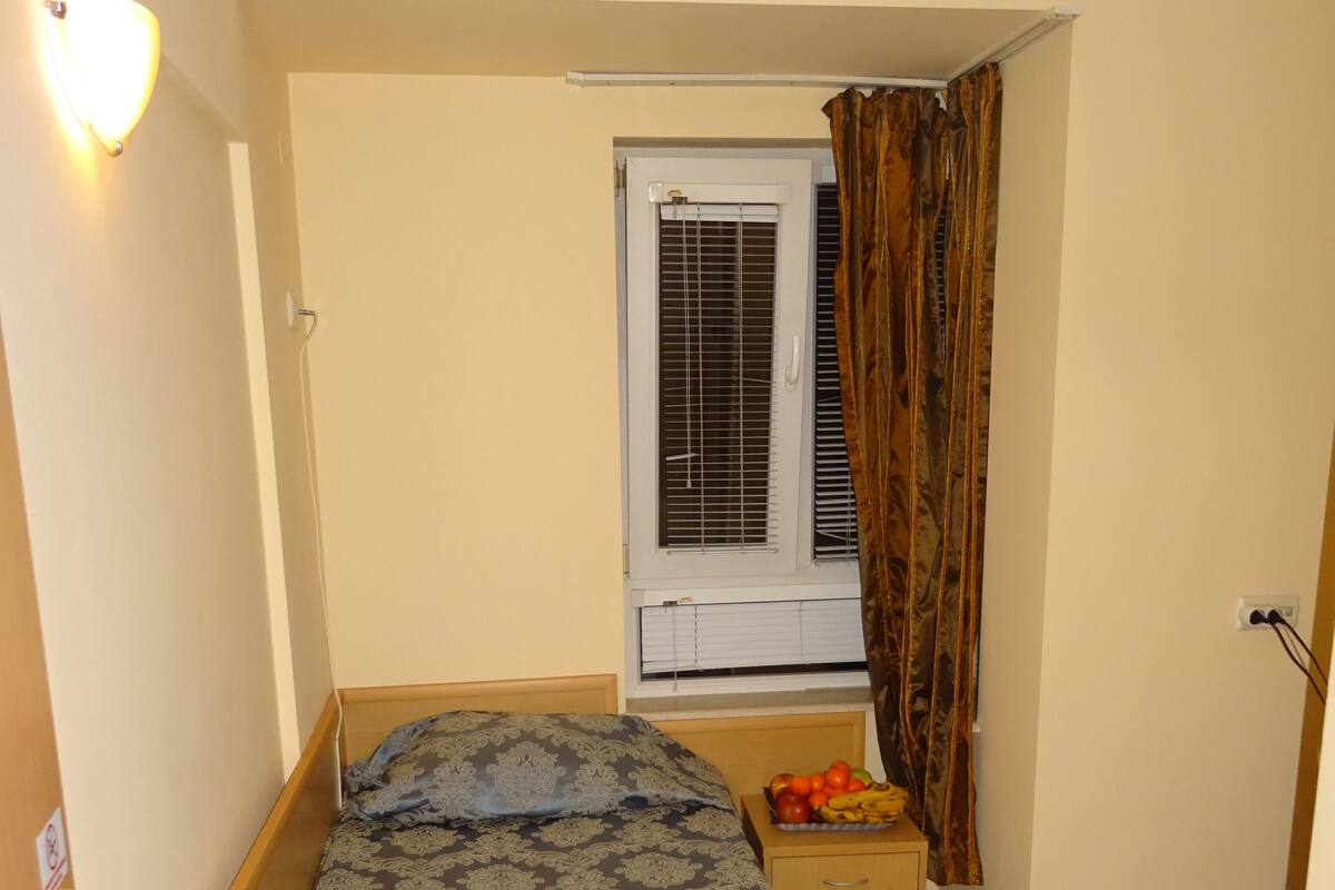 Una habitación pequeña y estrecha con una cama individual, una ventana con persianas, una mesita de noche con un frutero y una lámpara fijada a la pared.