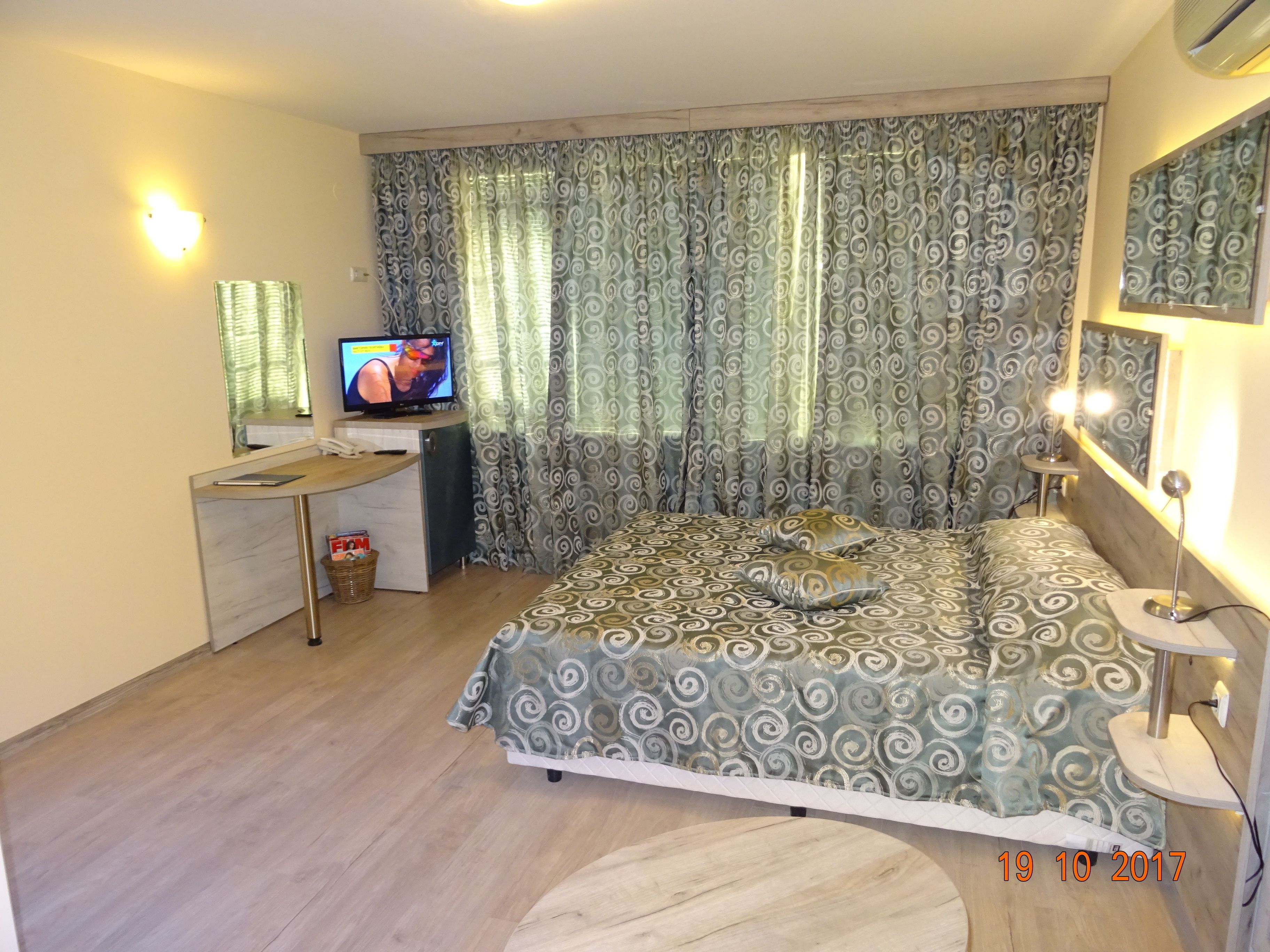 A bedroom with a bed, a desk with a television, a mirror, and curtains.