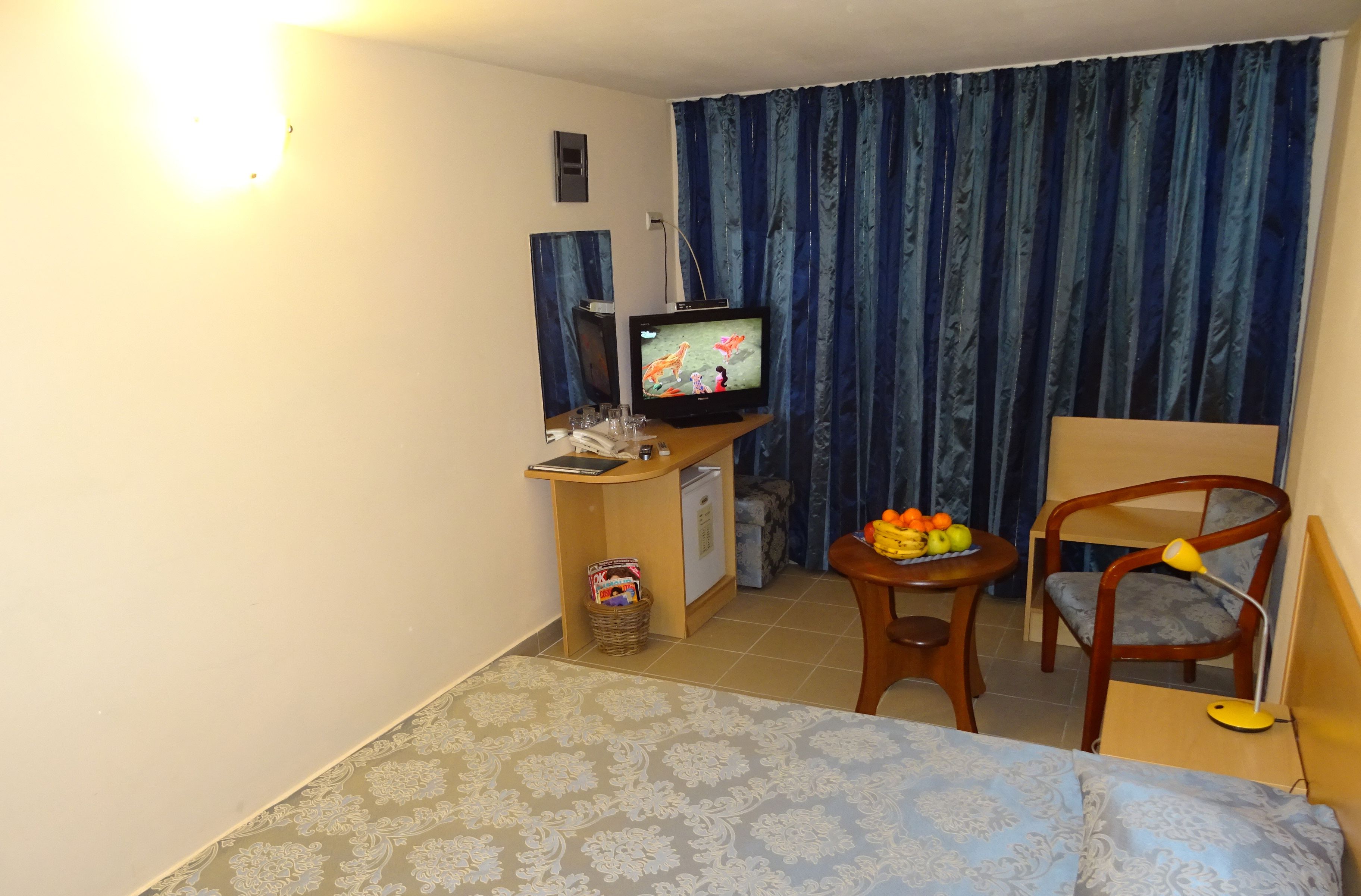 A compact hotel room with a bed, a small desk with a television, a chair, a table with fruit, and a lamp.