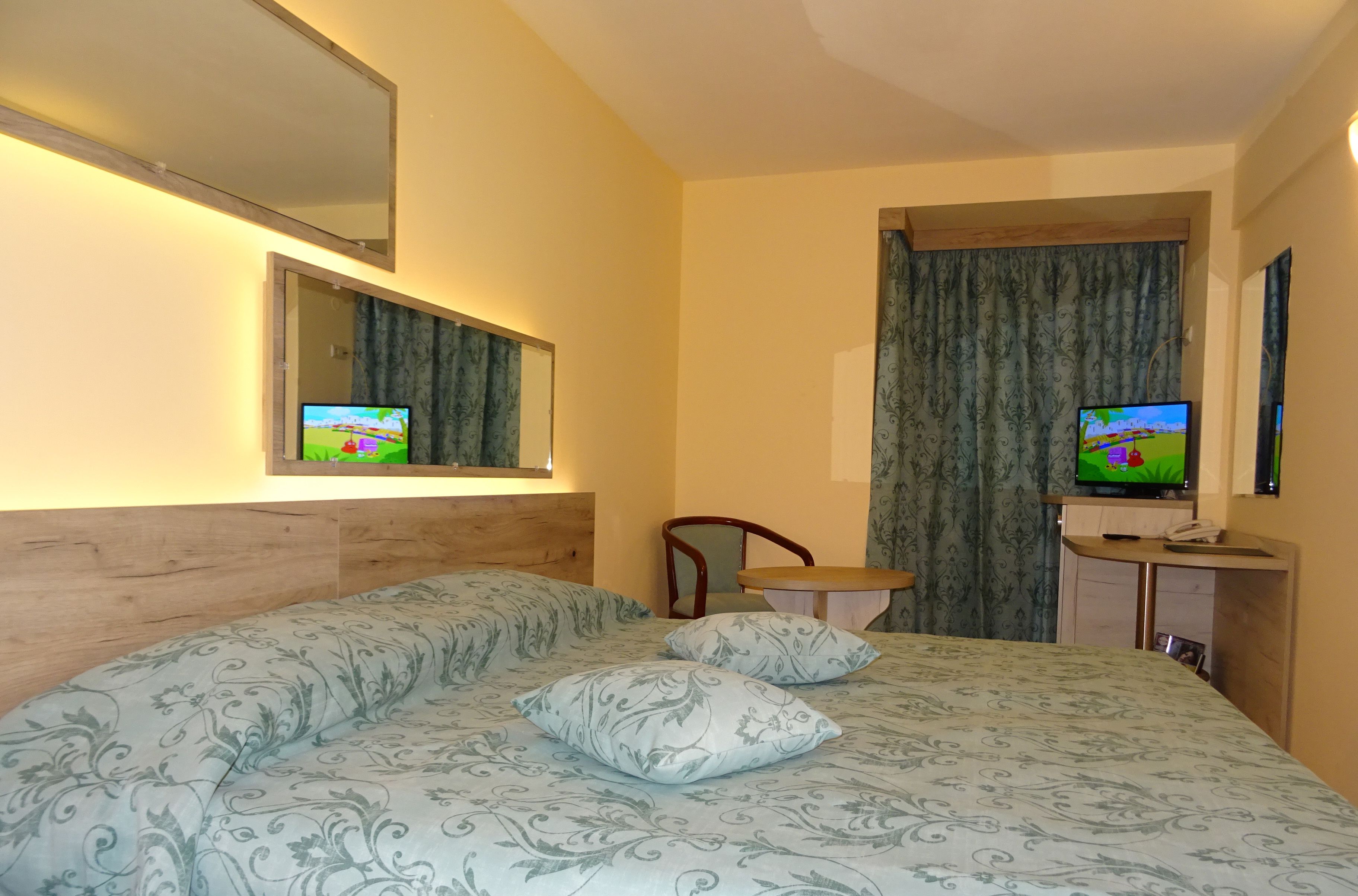 A hotel room with a large bed, two pillows, a wooden headboard, a bedside table with a lamp, a mirror, and a television on a stand.