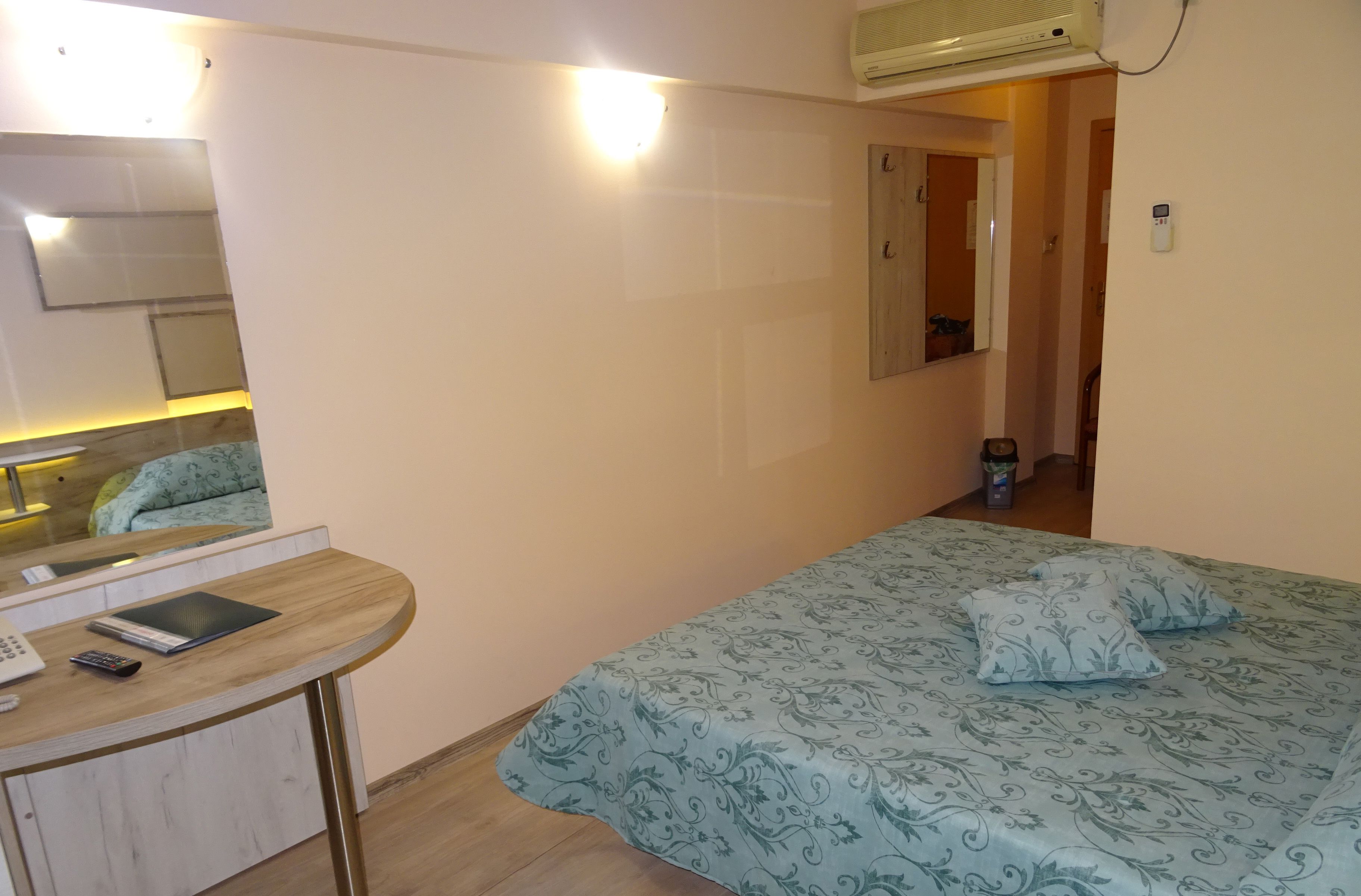 Un dormitorio con cama, escritorio con teléfono y tablet, espejo y puerta que da a otra habitación.