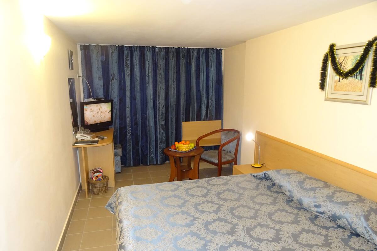 Una habitación de hotel con cama, escritorio con televisor, silla, mesita con fruta y cortina.