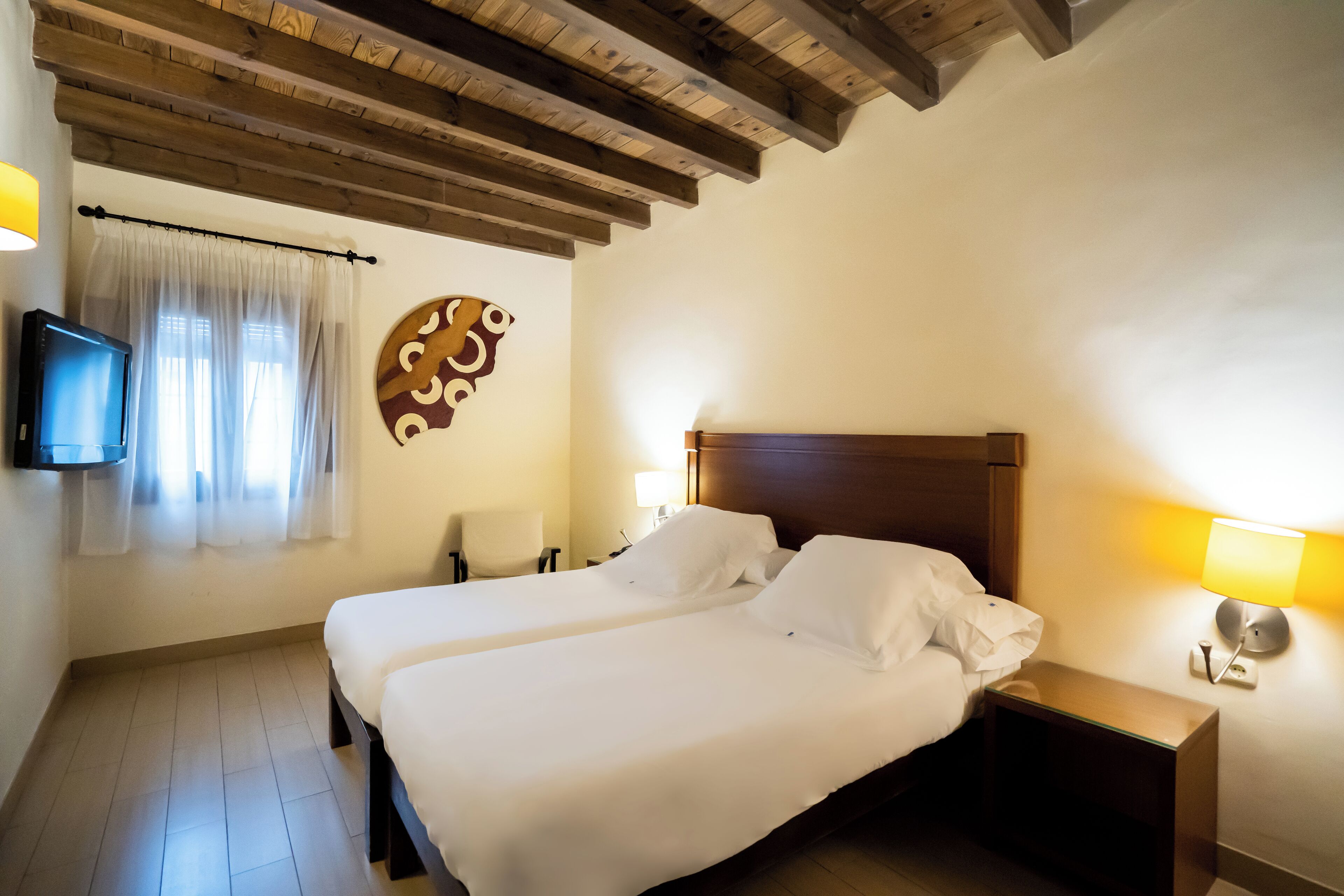Habitación de hotel con dos camas, vigas de madera en el techo, un televisor en la pared y una lámpara fijada a la pared.
