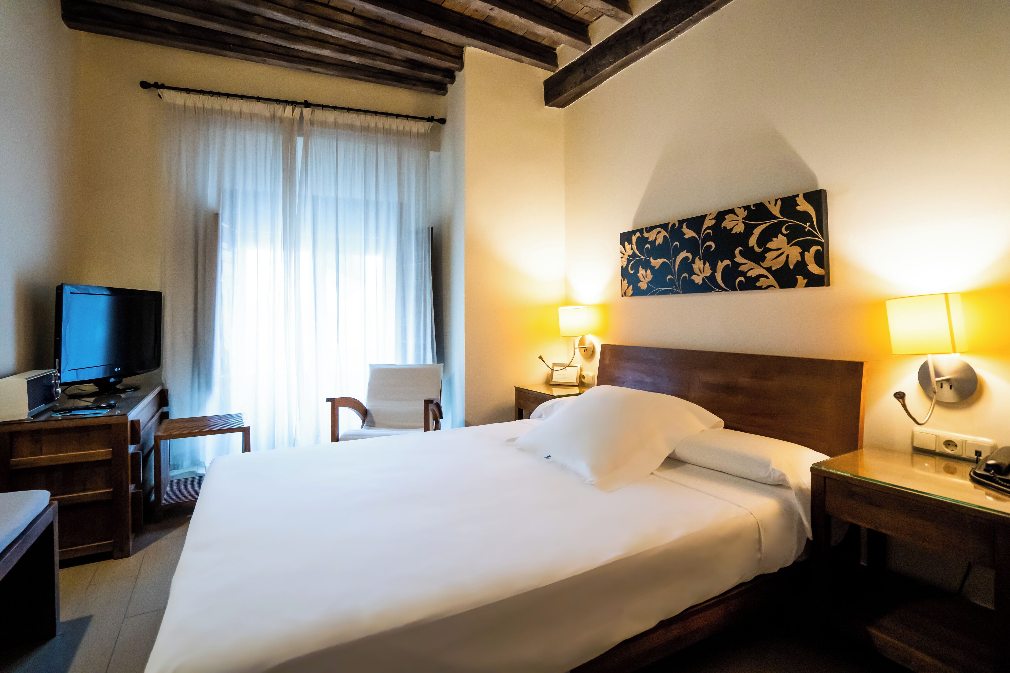 A hotel room with a large bed, a desk with a television, a chair, and two bedside lamps.