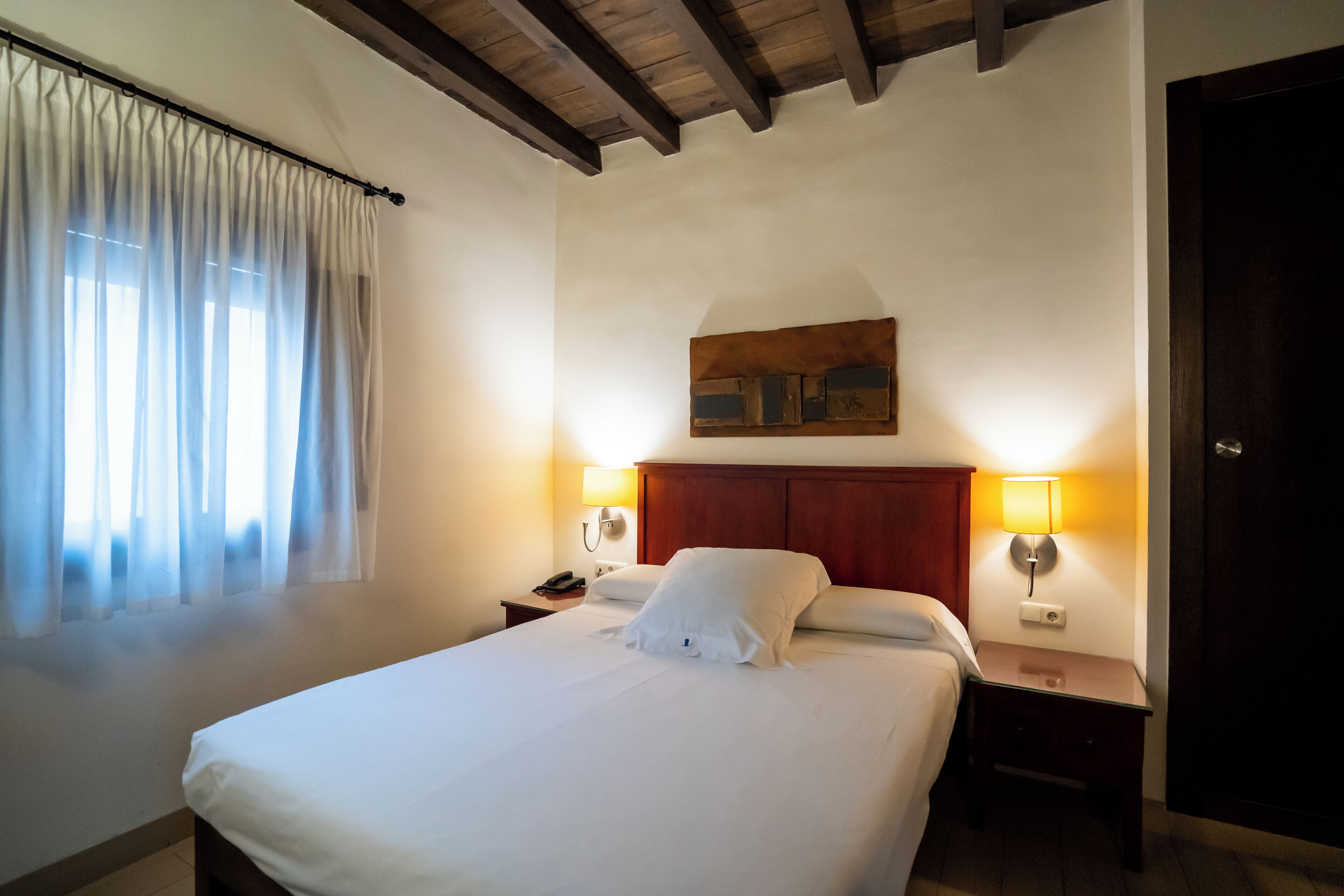 A hotel room with a large bed, two bedside tables with lamps, a wooden headboard, and a window with sheer curtains.
