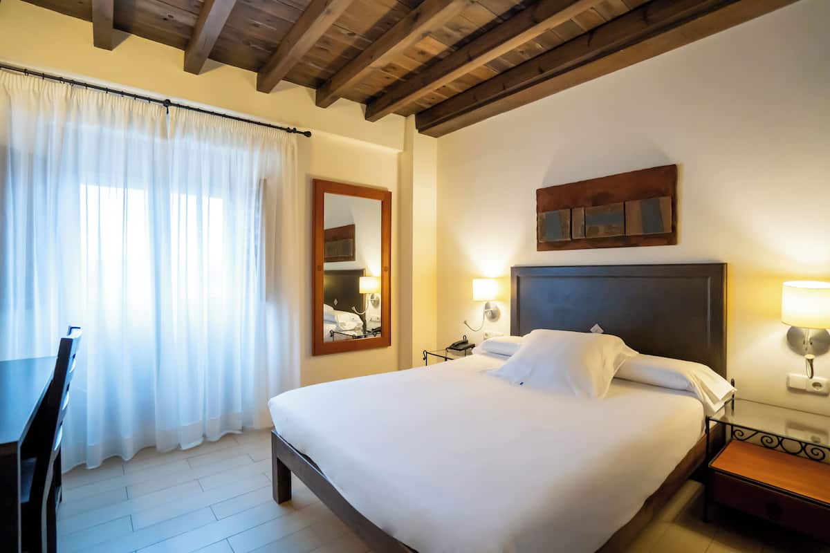Habitación de hotel con una cama grande, un escritorio y una silla. Cuenta con una ventana con cortinas, un espejo y una lámpara de pared.