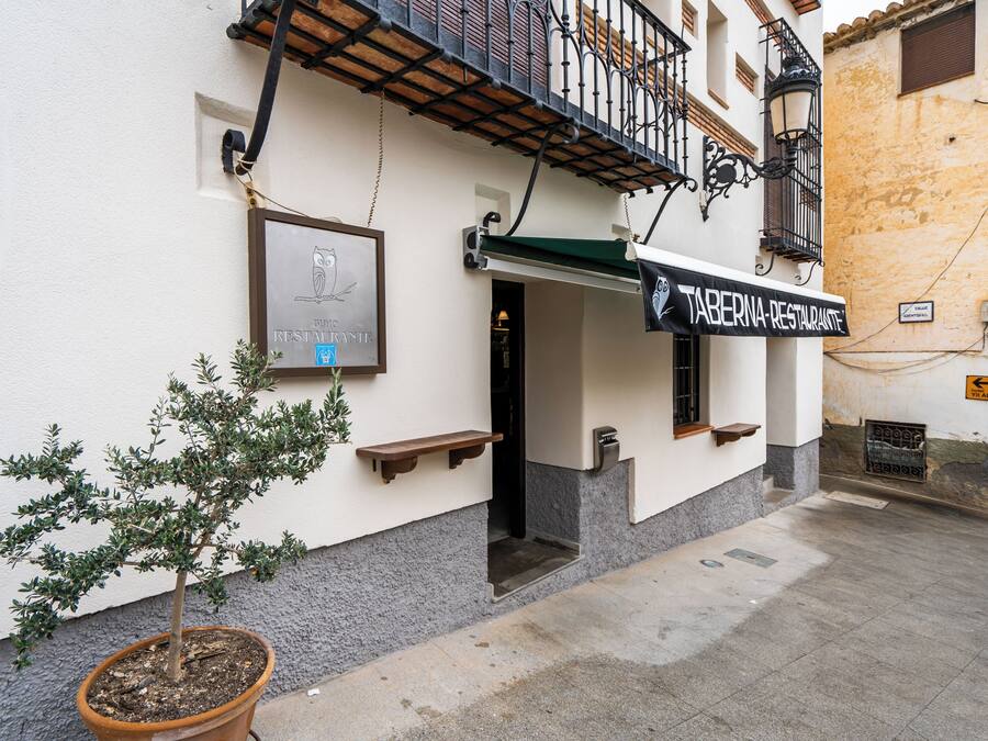Un edificio con un letrero de restaurante, una pequeña zona de mesas al aire libre y un árbol en maceta.