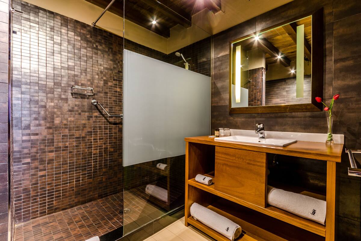 Un baño moderno con ducha envuelta en vidrio, tocador de madera con lavamanos y espejo con marco de madera.