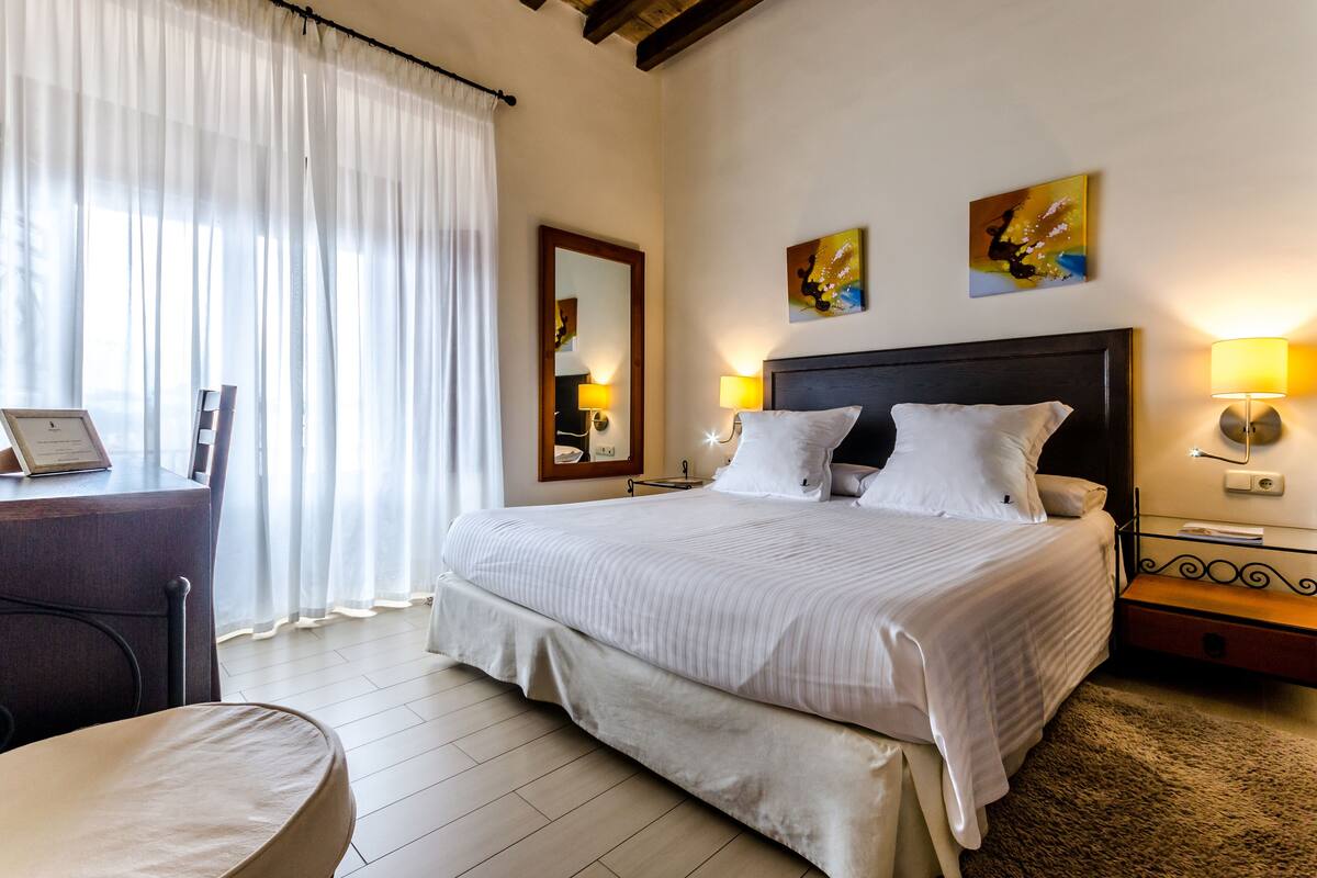 Habitación de hotel con una cama grande, un escritorio con silla, una mesita pequeña y dos lámparas de pared.