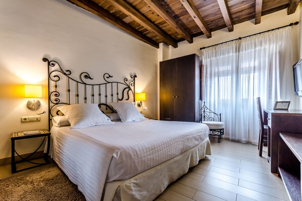 A neatly made bed with a headboard, bedside lamps, a wooden desk, and a wardrobe in a well-lit room with a tiled floor.