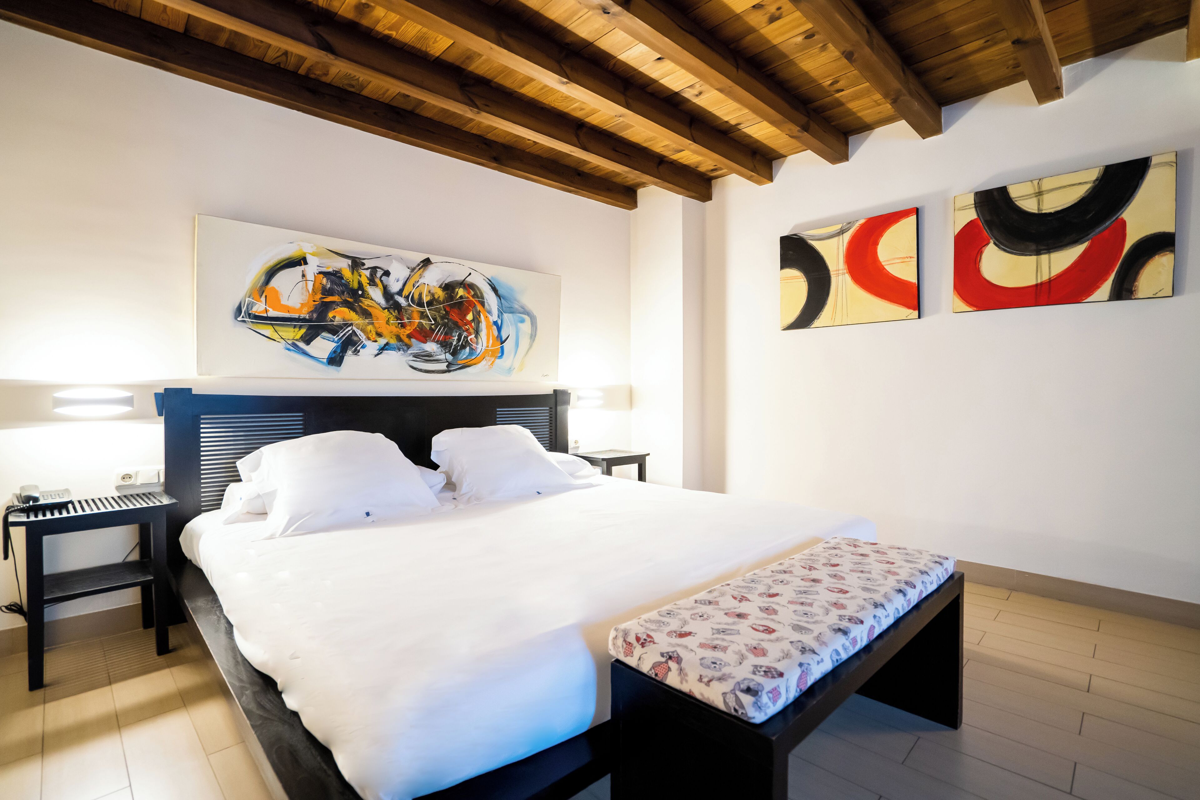 Une chambre à coucher comprenant un lit, des tables de chevet, un banc et des œuvres d’art abstraites accrochées au mur.