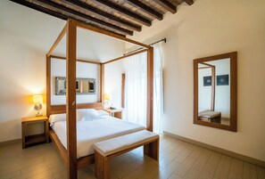 Uma cama com dossel, um banco de madeira, mesas de cabeceira com abajures, um espelho grande e vista do quarto.
