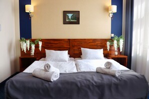 A hotel room with a wooden headboard, two white pillows, a dark bedspread, and two bedside tables with lamps and flower arrangements.