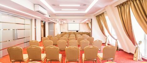 Uma sala de conferências com fileiras de cadeiras, uma tela de projetor e um grande janelão com cortinas.