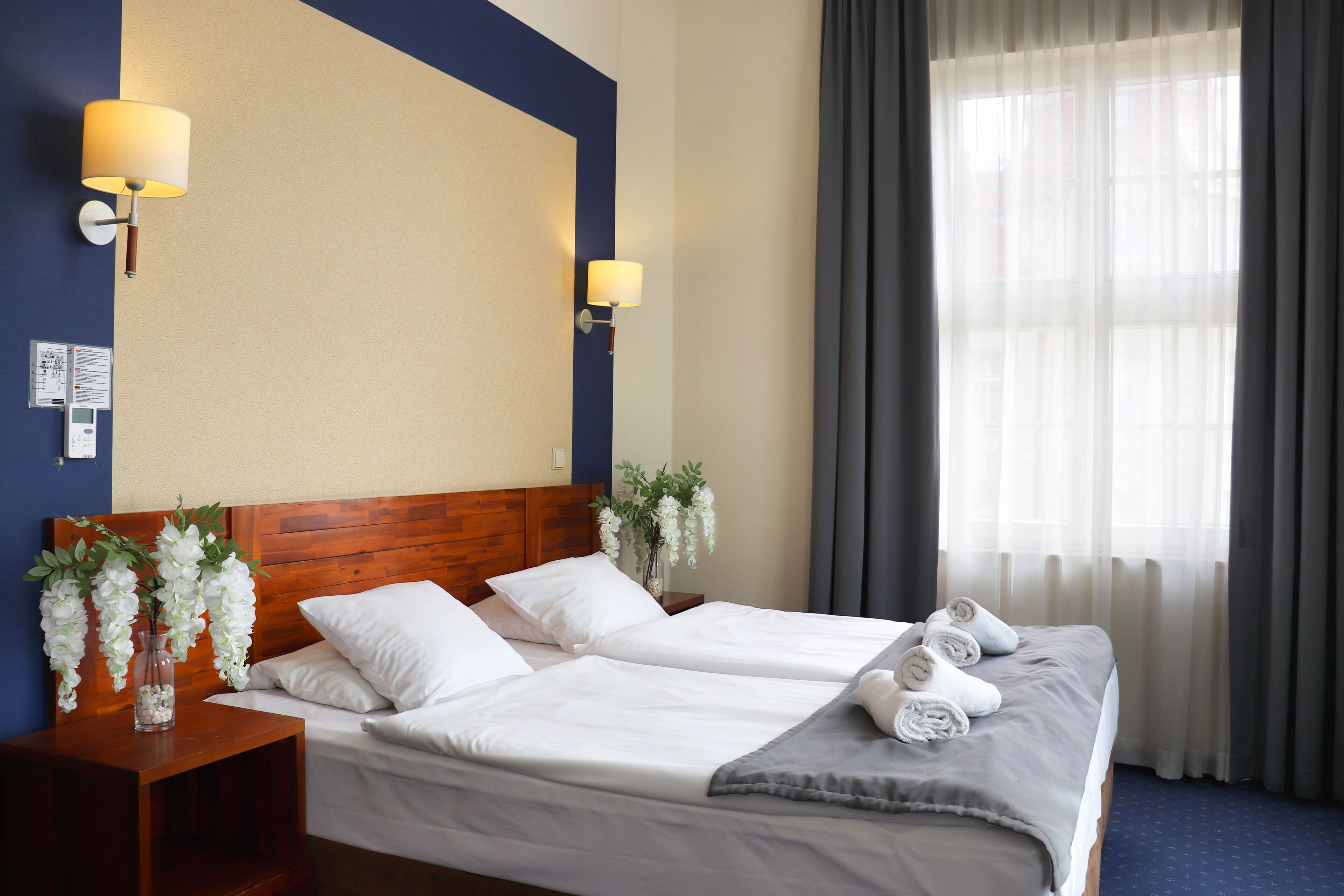 A hotel room with two beds, a wooden headboard, white bedding, a vase with white flowers, and a window with curtains.