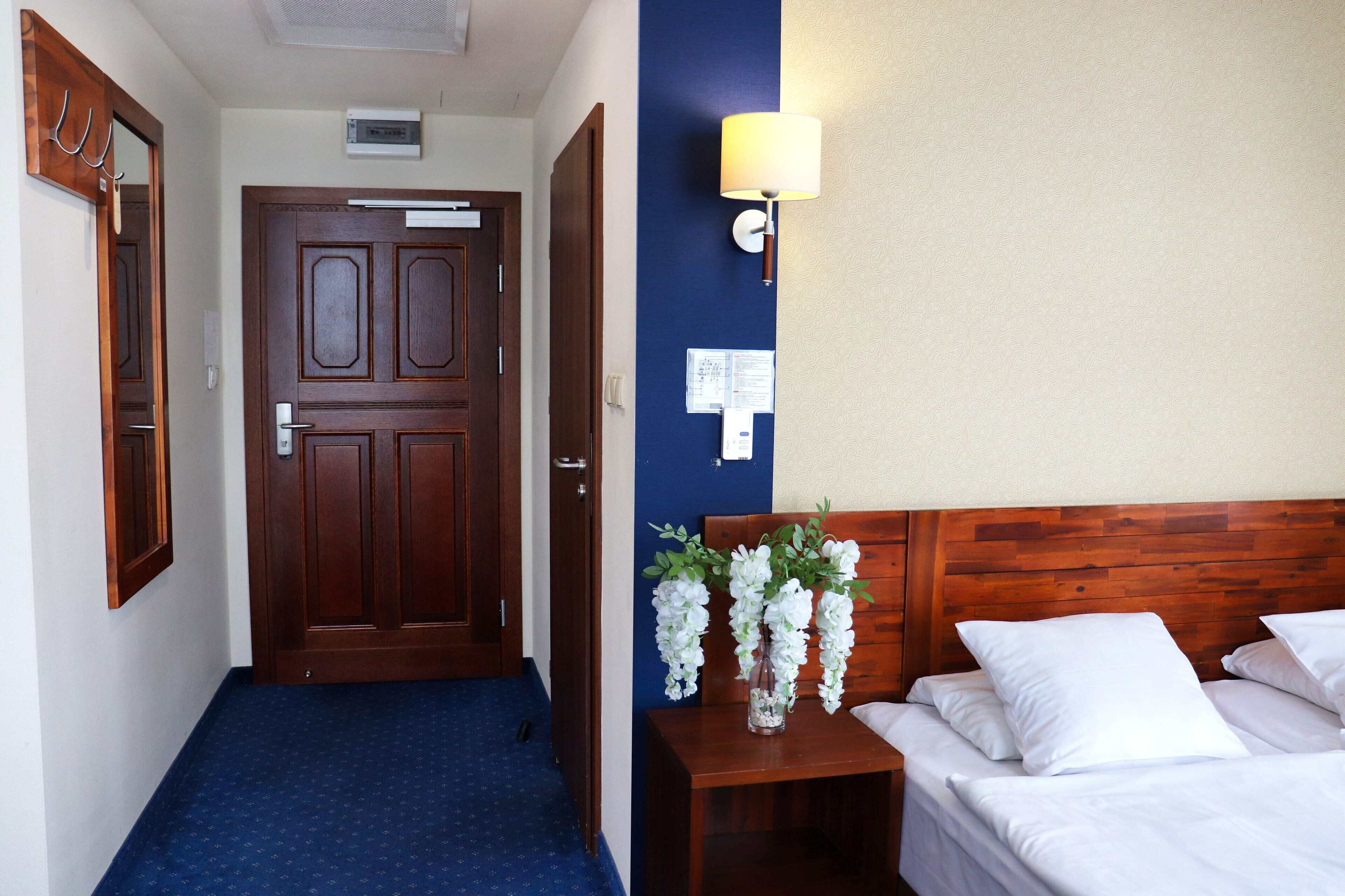 A hotel room with a wooden door, a blue carpeted hallway, a bedside table with a vase of flowers, and a bed with white bedding.