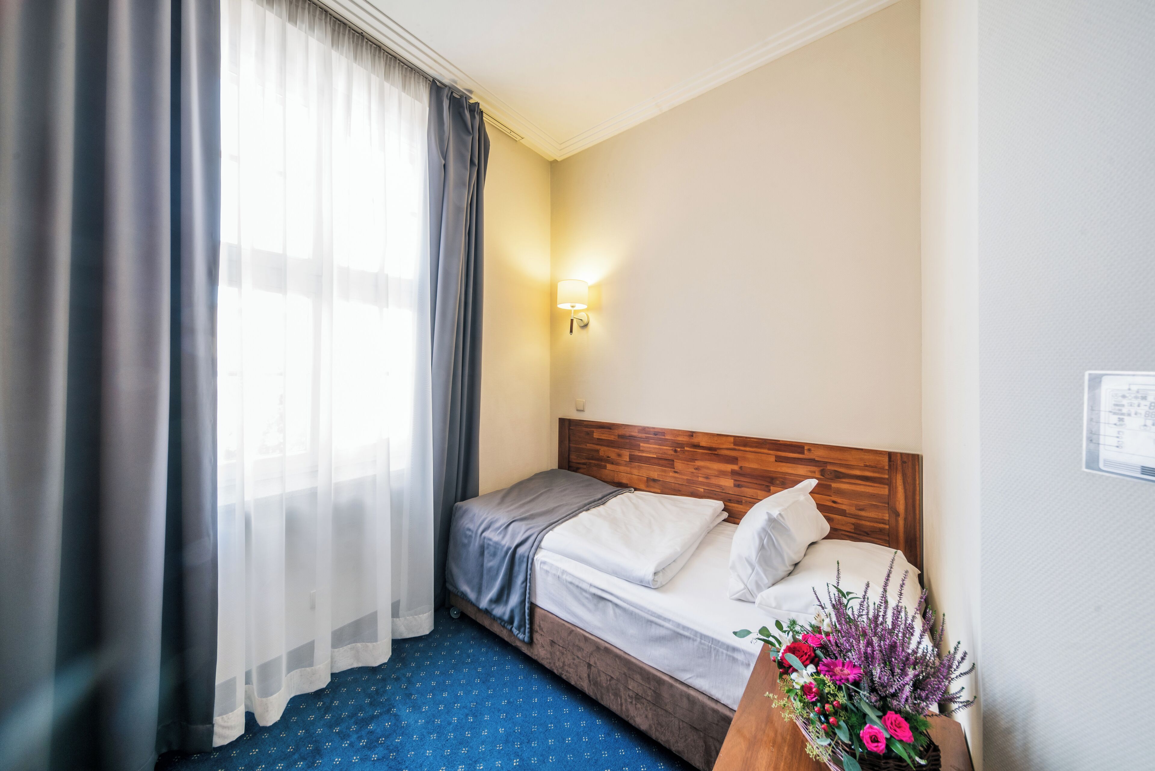 Una habitación de hotel con una cama, una mesita de noche con lámpara, una ventana con cortinas y una luz instalada en la pared.