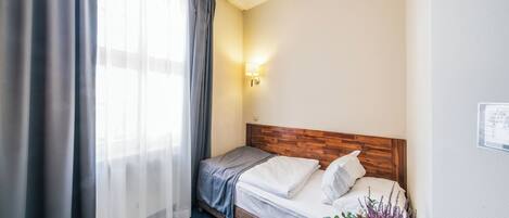 A hotel room with a bed, a nightstand with a lamp, a window with curtains, and a wall-mounted light.