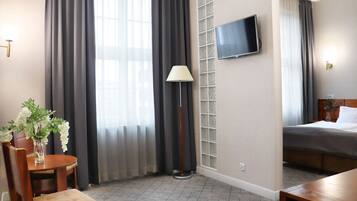 Habitación de hotel con una cama, un televisor montado en la pared, una lámpara, una silla y una mesita con un jarrón de flores.