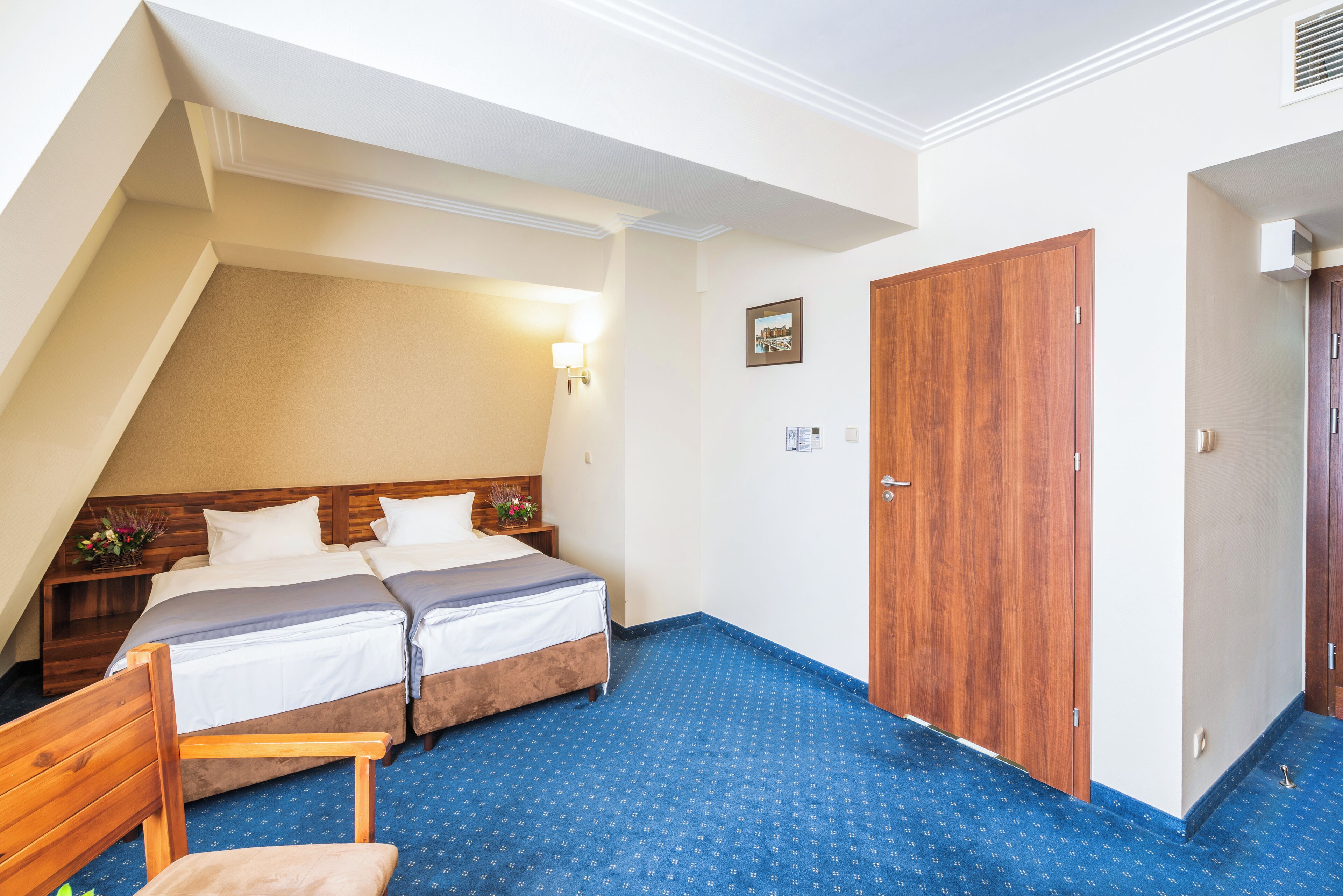 Una habitación con dos camas, un armario de madera, una mesita de noche con lámpara y una alfombra azul estampada.