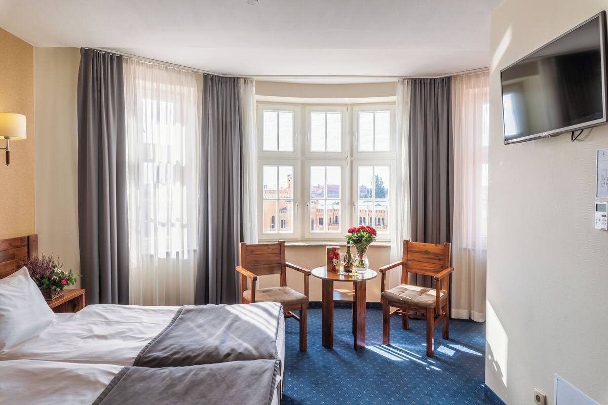 A hotel room with a bed, a desk with a chair, a TV, and a window with curtains.