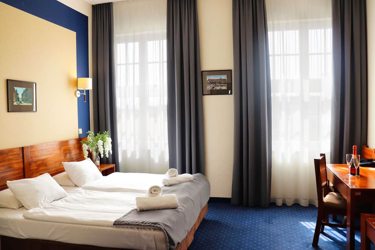 A hotel room with two beds, a wooden headboard, a dining table with chairs, and large windows with curtains.
