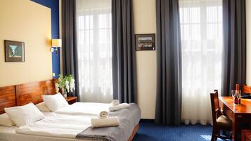 Habitación de hotel con dos camas, cabecera de madera, una mesa de comedor con sillas y ventanas grandes con cortinas.