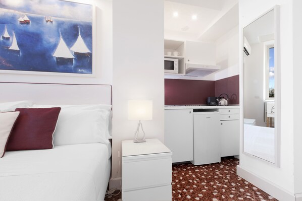 Un dormitorio con cama, mesita de noche, un cuadro, una zona de cocina con armarios y microondas, y un baño con espejo y lavamanos.