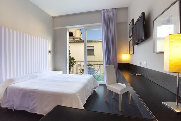 Habitación de hotel con cama, escritorio, silla, televisión y balcón con mesa y sillas.