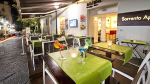 Un área de comedor al aire libre con mesas cubiertas con manteles verdes, sillas y un cartel de menú en la pared.