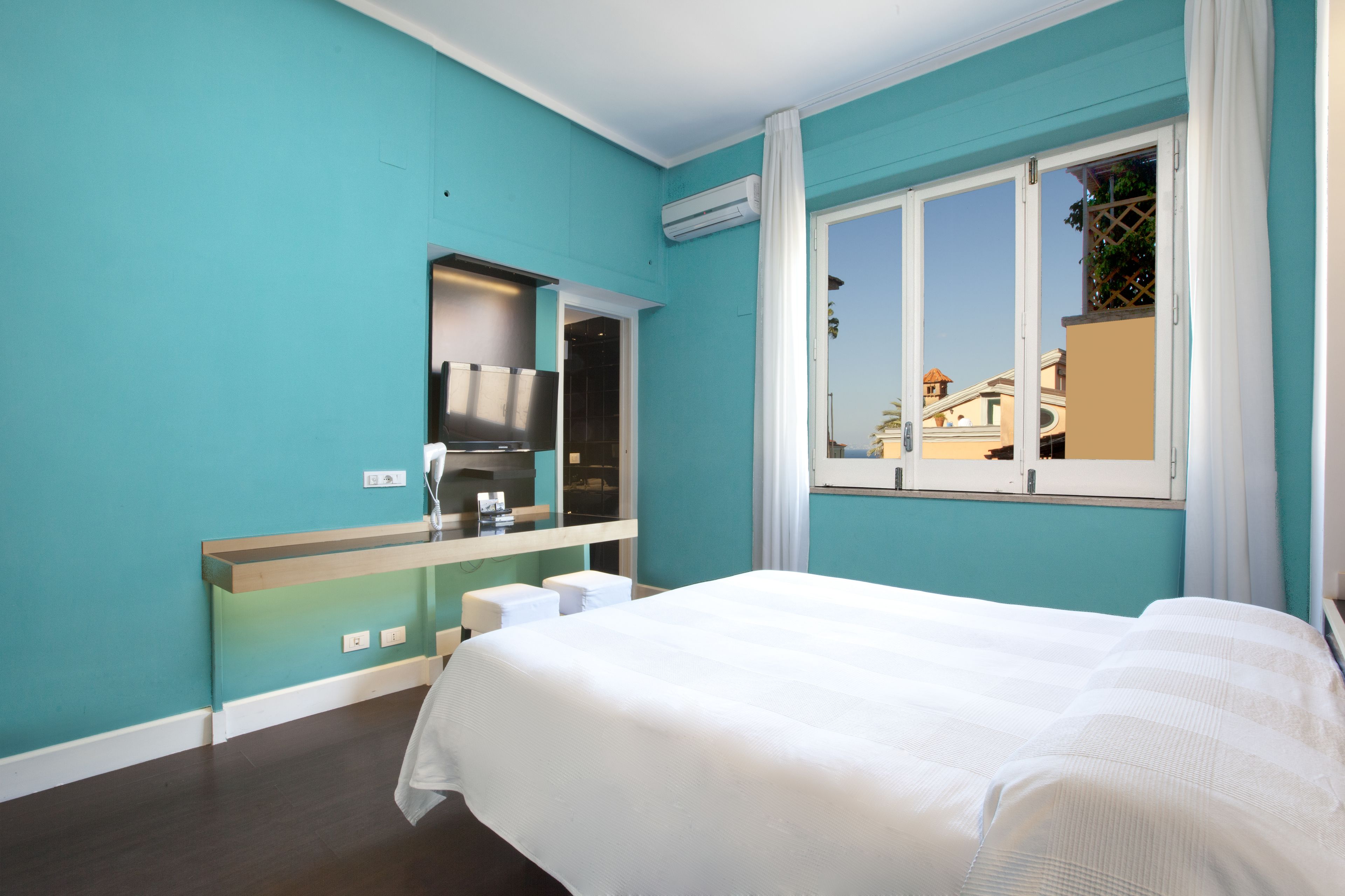 A hotel room with teal walls, a large bed, a built-in TV, and a window with a view of buildings.