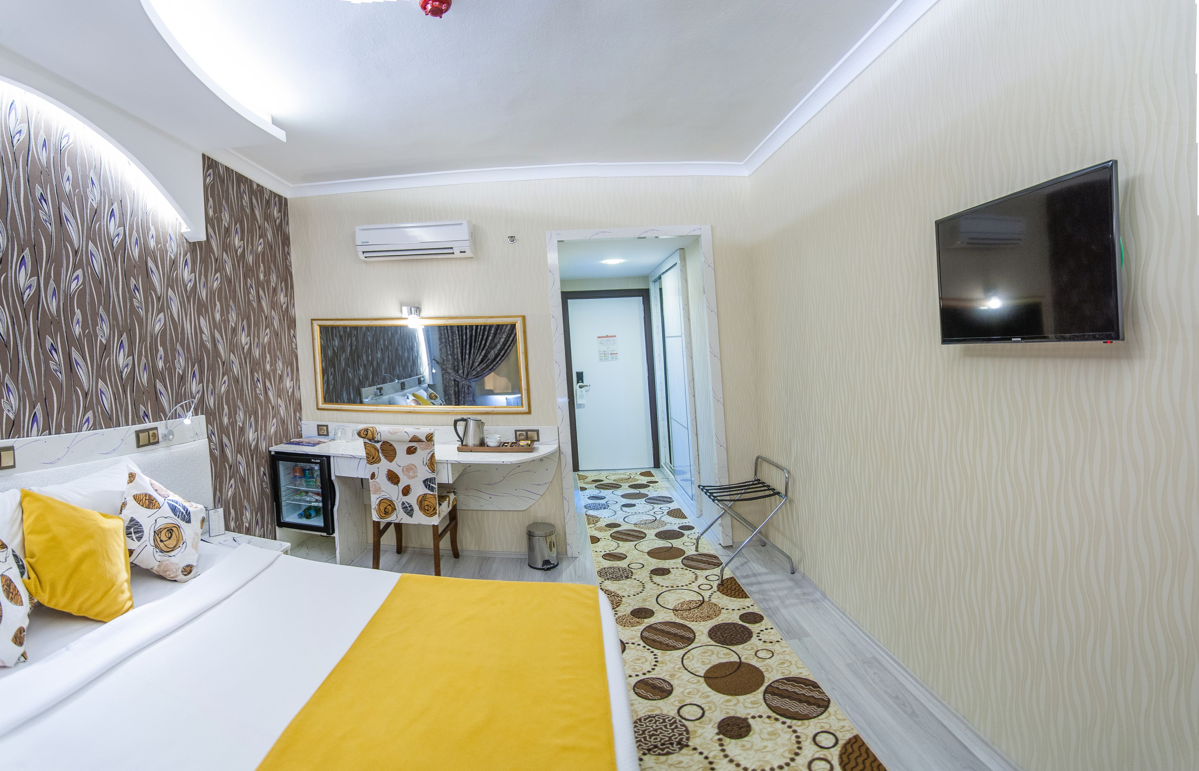 Habitación de hotel con cama, escritorio, espejo, televisor y baño visible a través de una puerta abierta.