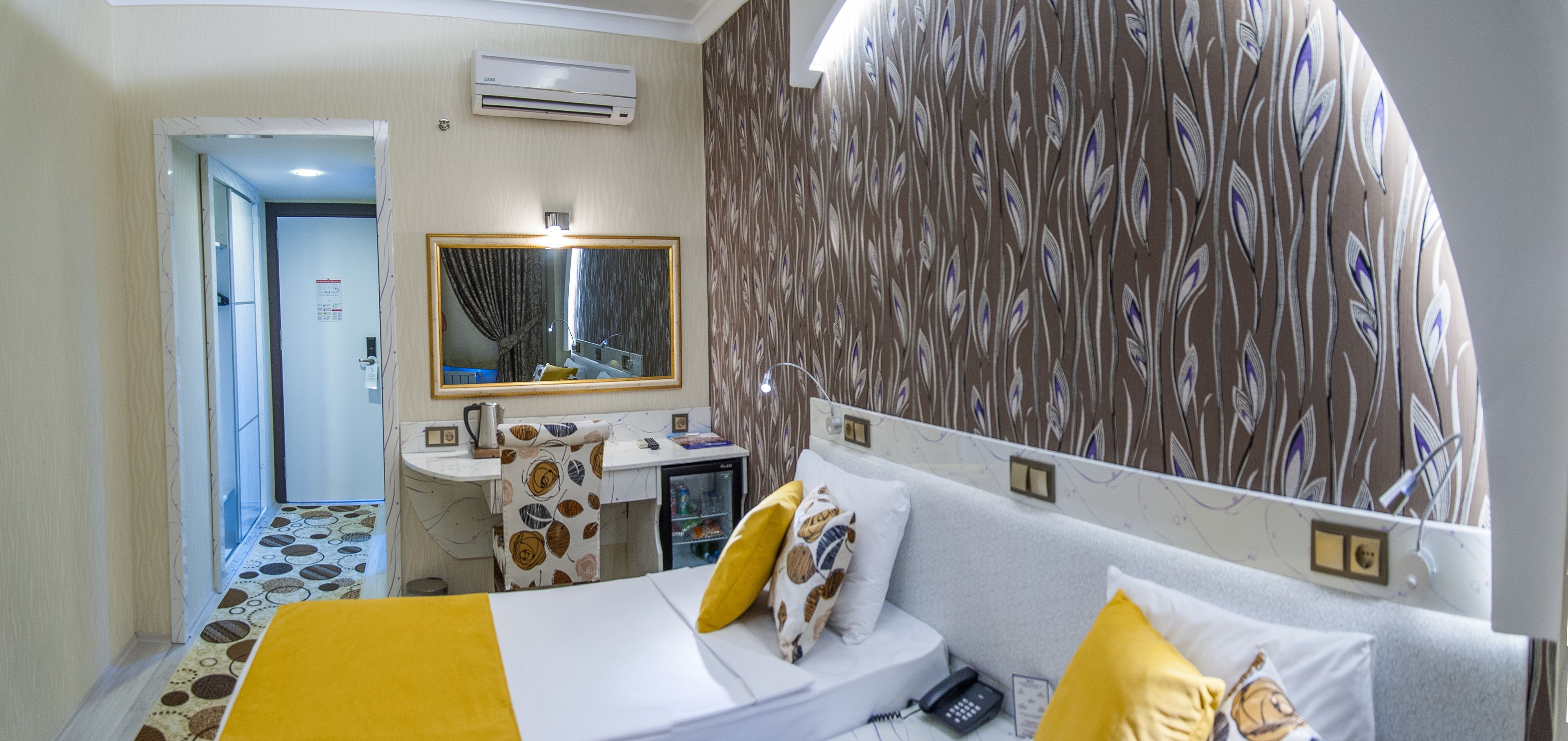 A hotel room with a bed, bedside table, and a desk with a chair. There is a mirror above the desk, and a wall with a leaf pattern.