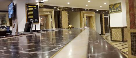 Zona de recepción de un hotel con mostrador de mármol, señal de ascensor y señalización de cuarto de equipaje.