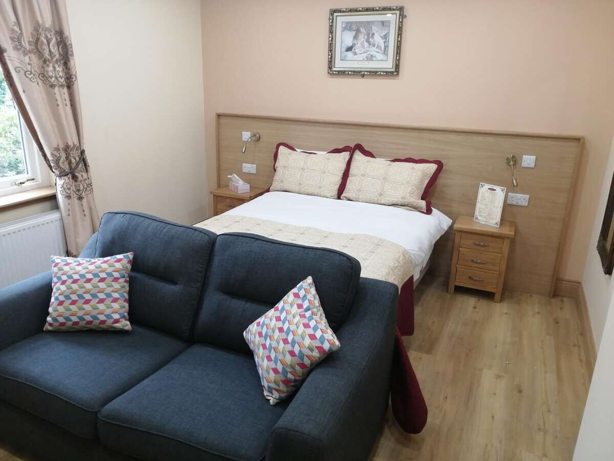 Quarto com cama, cabeceira de madeira, mesa de cabeceira, sofá azul e duas almofadas estampadas.