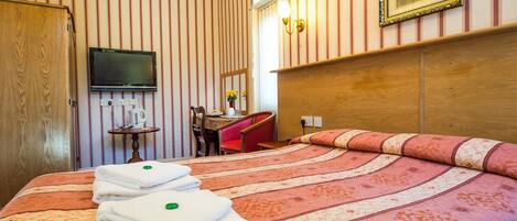 A hotel room with a bed, a TV mounted on the wall, a desk with a chair, and a wooden wardrobe.
