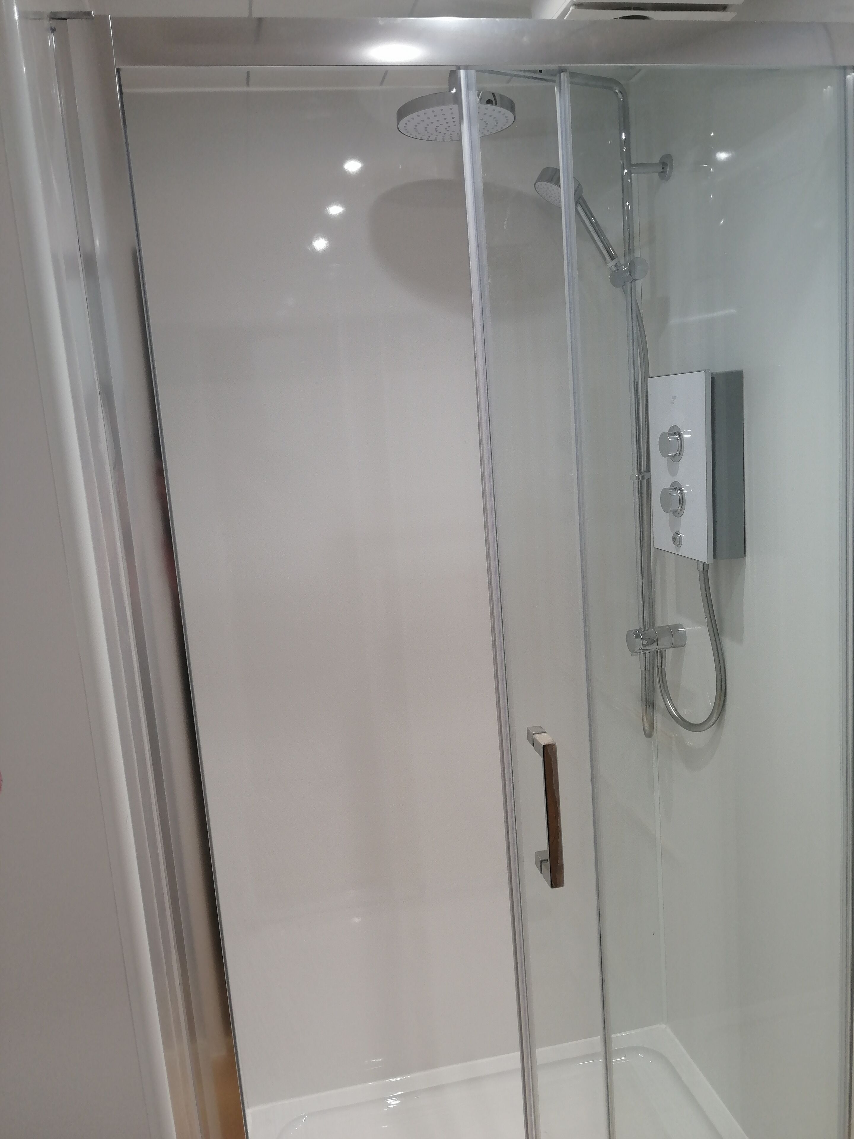 Un espace douche entièrement vitré, doté d’un pommeau de douche à main et d’un panneau de commande fixé au mur.