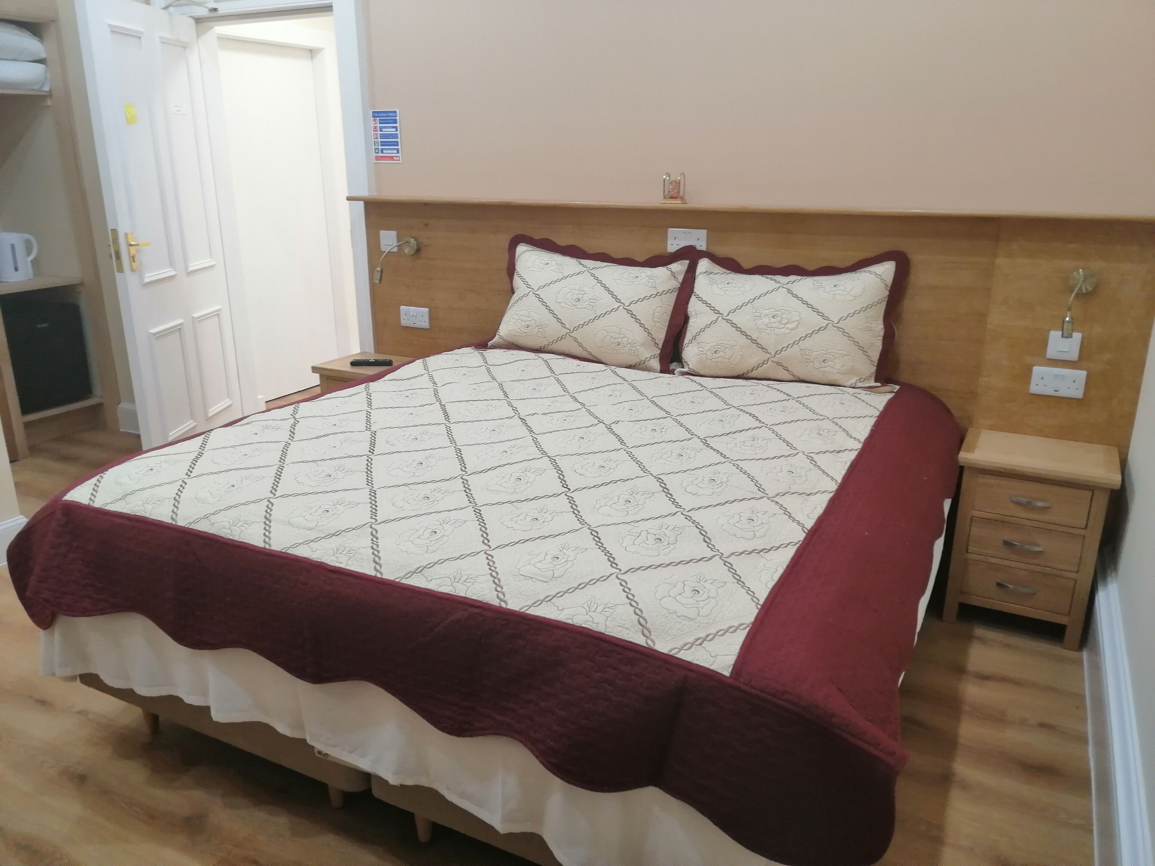A neatly made bed with a patterned quilt and a red throw at the foot. There is a wooden nightstand with a lamp and a small drawer.