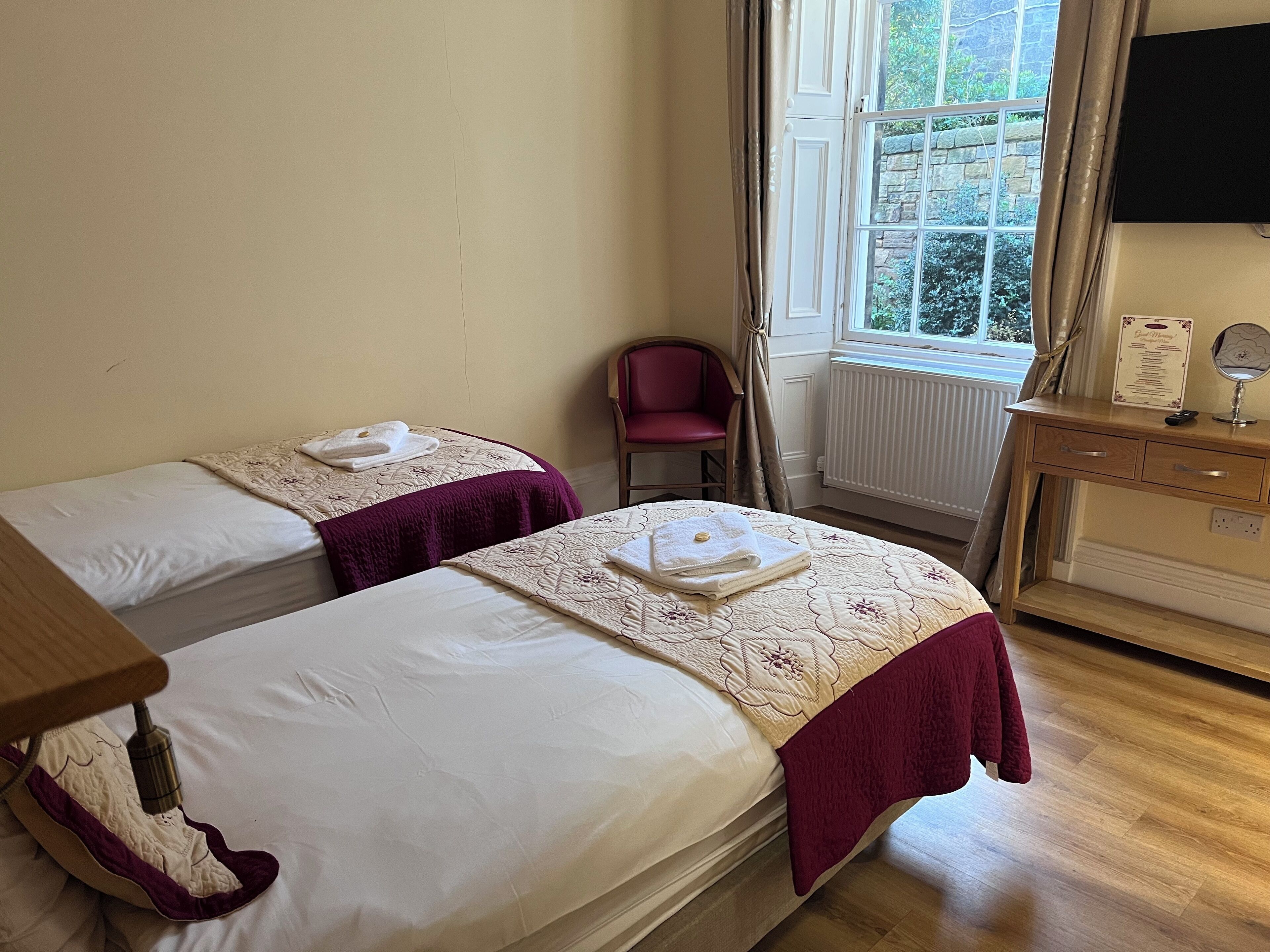 Una camera d'albergo con due letti, una scrivania in legno, una sedia e una finestra con tende.