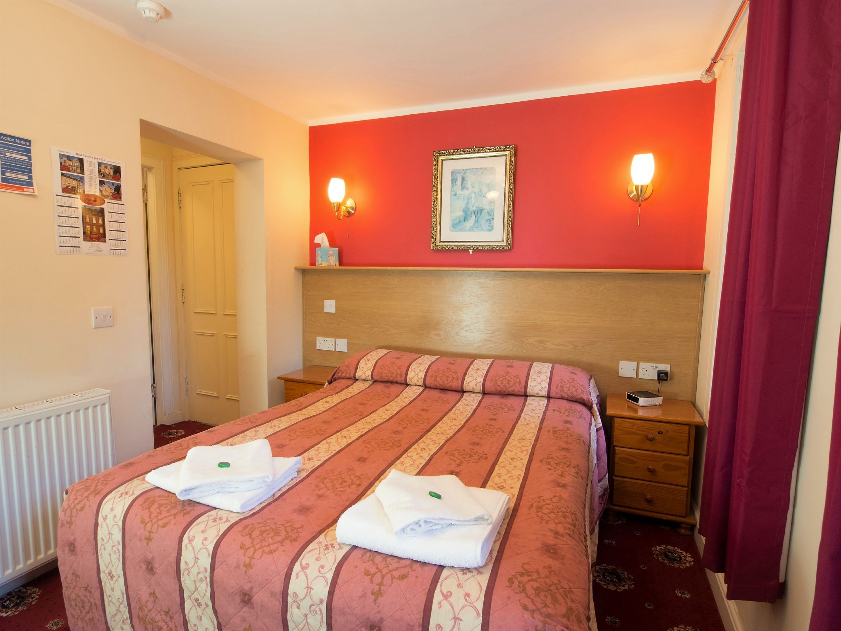 A hotel room with a bed, two bedside tables, a nightstand, a radiator, and a painting on the wall.