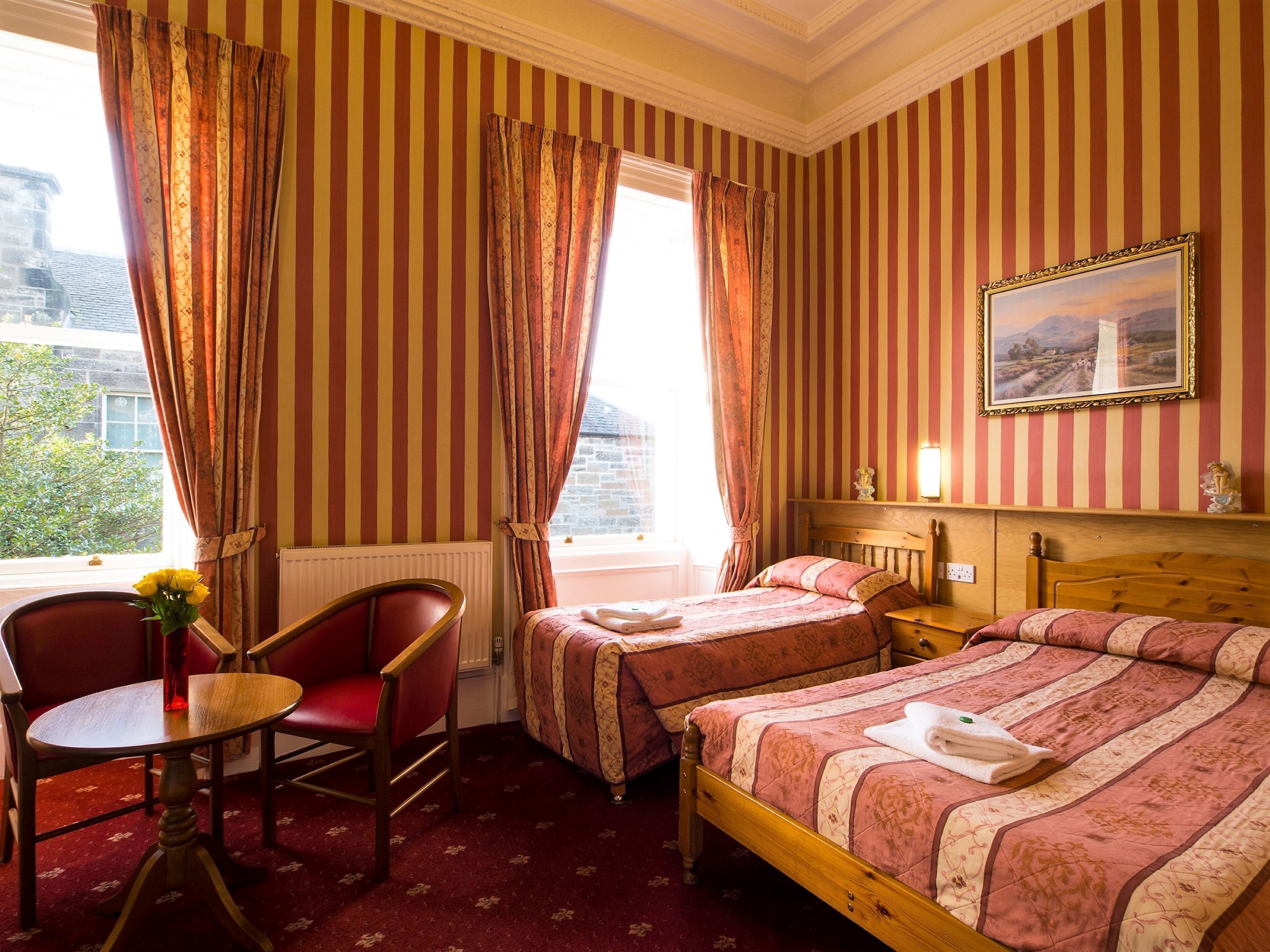 Ein Hotelzimmer mit zwei Betten, einem Holz-Kopfteil, einem kleinen Tisch mit Blumenvase und einem Fenster mit Vorhängen.