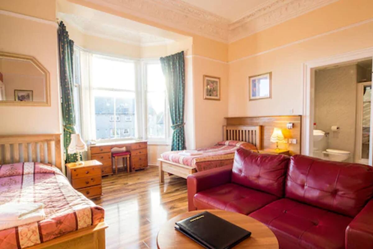 A hotel room with a bed, a red leather sofa, a wooden table, a desk, and a window with curtains.