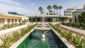 Un patio con una fuente central, rodeado de palmeras y un edificio con techo verde.
