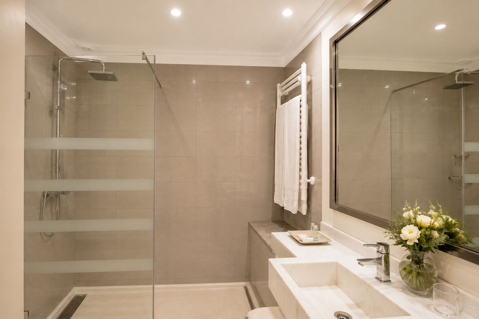 A modern bathroom with a glass-enclosed shower, a large mirror, and a vanity with a sink and a vase of flowers.