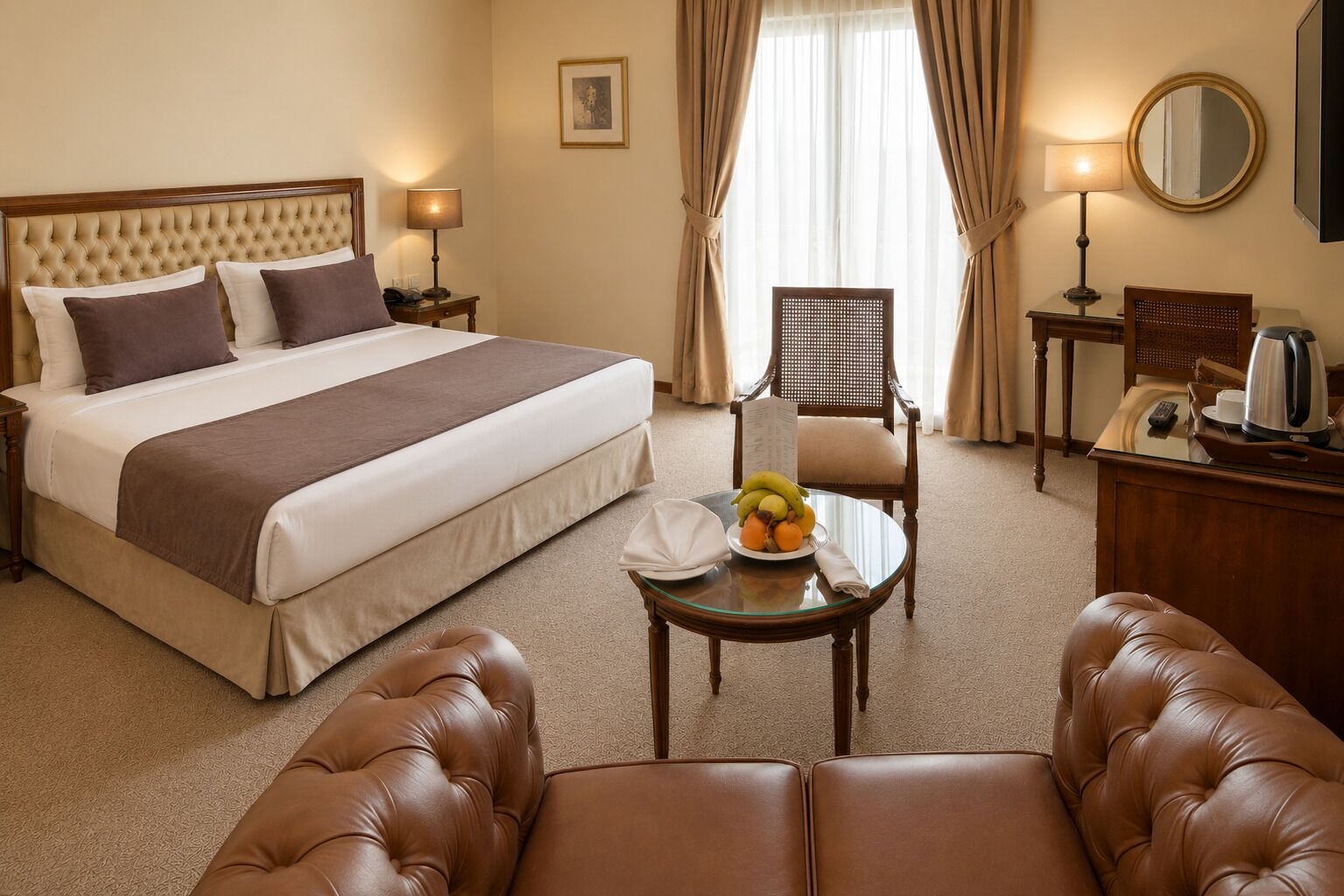 Een hotelkamer met een groot bed, een eettafel met fruit, een leren fauteuil, een houten bureau en een spiegel.