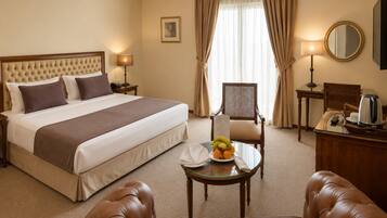 Een hotelkamer met een groot bed, een eettafel met fruit, een leren fauteuil, een houten bureau en een spiegel.