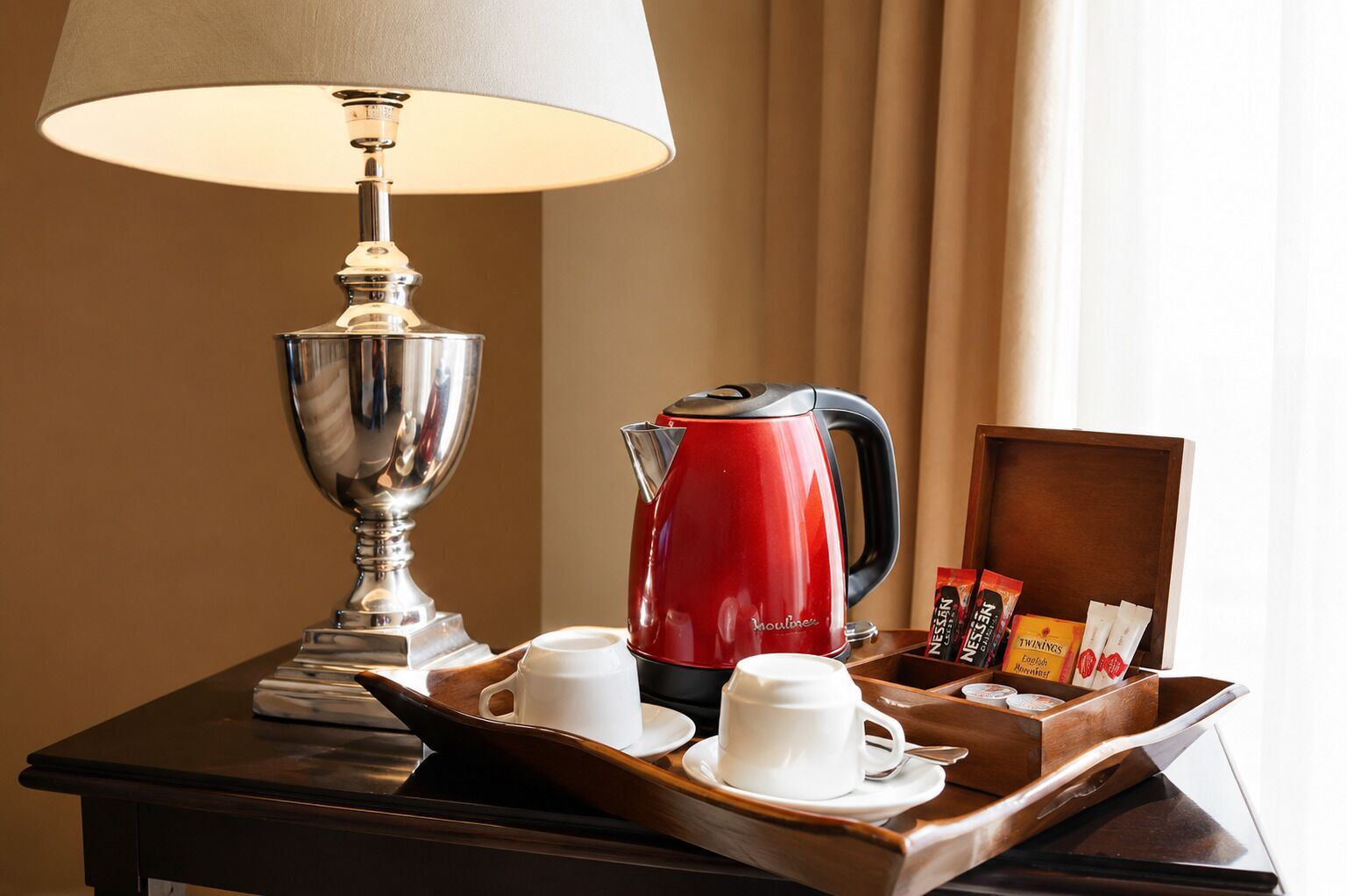 Mesinha de cabeceira em quarto de hotel, com uma lâmpada, uma chaleira elétrica vermelha, duas chávenas brancas, um tabuleiro com saquinhos de chá e uma caixa de madeira.
