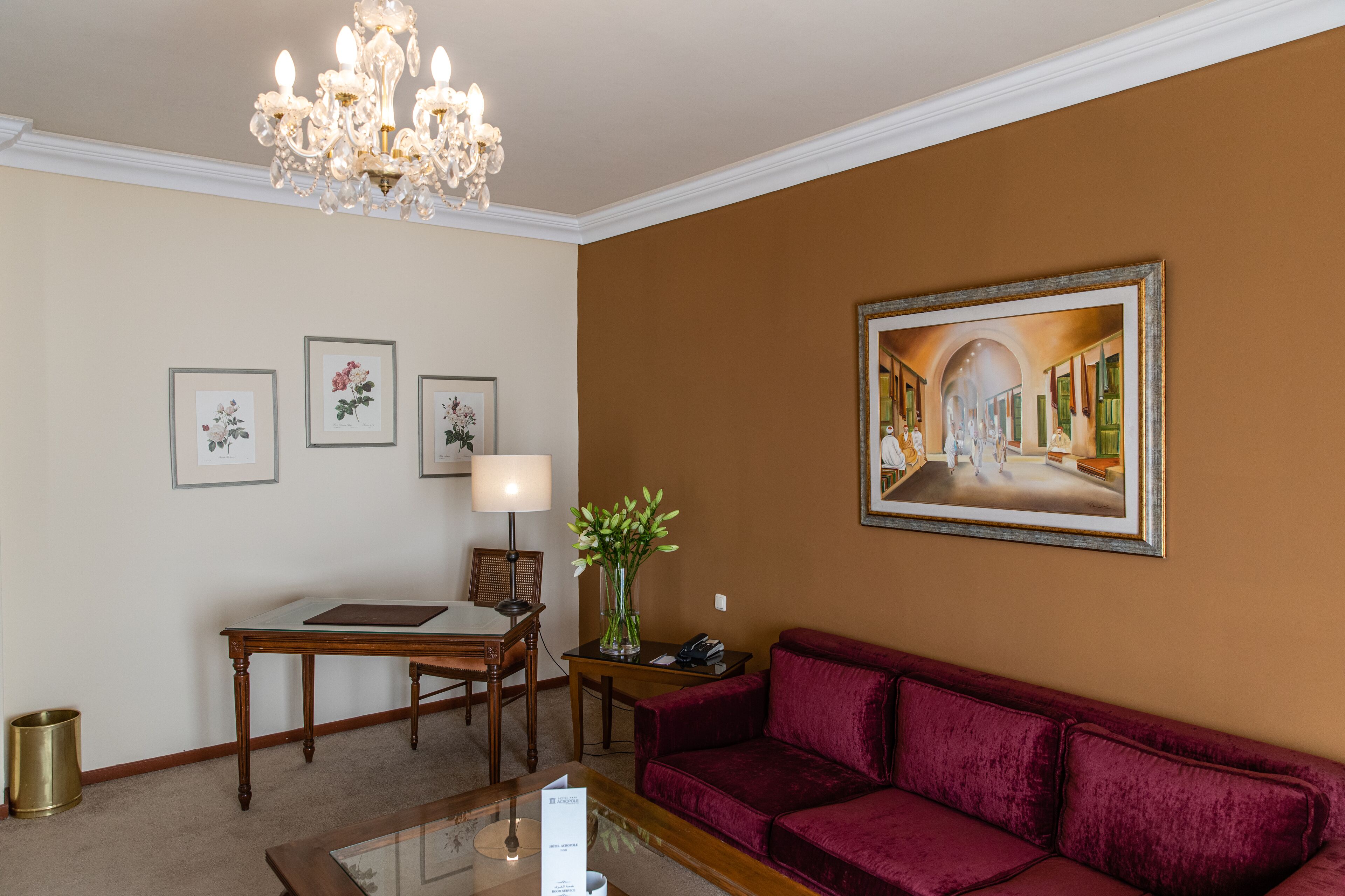 Habitación de hotel con sofá de terciopelo rojo, una mesa de madera, un candelabro, cuadros con motivos florales y un cuadro de un pasillo.