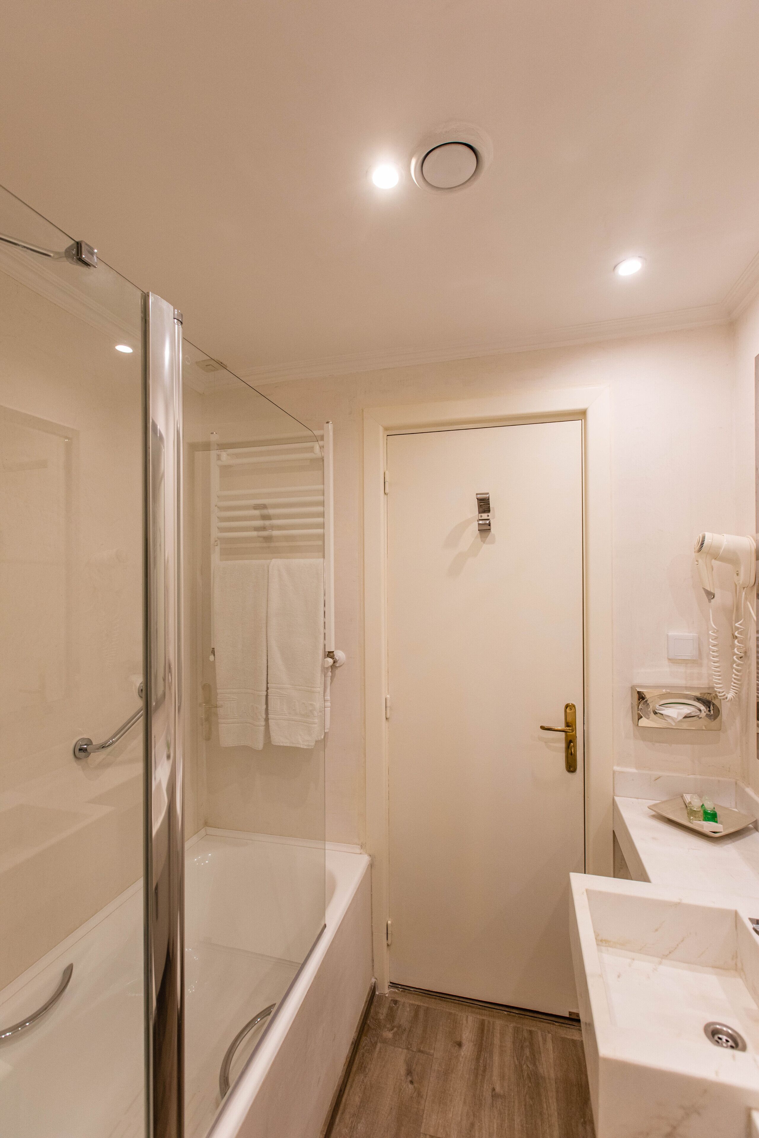 Un baño con ducha envuelta en vidrio, una puerta blanca, un lavabo con espejo y un secador de pelo en la pared.
